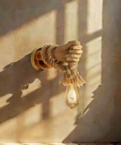 Une pièce murale de style vintage, connue sous le nom de *Lampe Murale Vintage Style Robot avec Corde de Chanvre E27*, présente une sculpture en forme de main tenant une ampoule suspendue entrelacée dans une corde de chanvre. La lumière du soleil projette de douces ombres sur le mur, créant un jeu artistique d'ombre et de lumière.