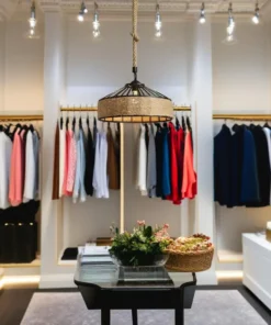 Une boutique de vêtements chic est dotée d'une table centrale en bois décorée de plantes. Une variété de vêtements colorés, tels que des vestes, des chemises et des robes, sont élégamment exposés sur des portants dorés étincelants le long des murs. Des spots de plafond vintage de la gamme Lustre vintage rustique CORDA Noir beige (diam. 30cm E27) illuminent chaleureusement la scène.